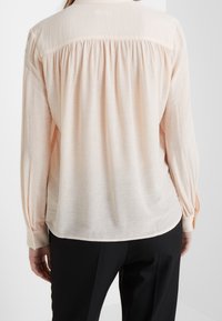 Hedvábná dlouhá blůza v broskvové barvě s nabraným zadním dílem, jemnou texturou, knoflíkovými manžetami a kulatým lemem, v kombinaci s černými kalhotami.