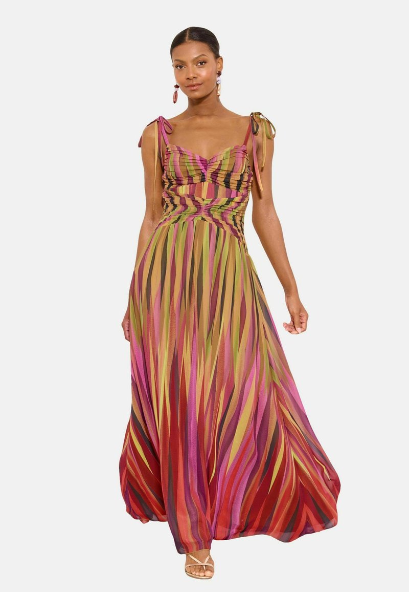 Woman wearing a sleeveless, multicolored striped maxi dress with tied shoulder straps and sandals, standing against a plain background.