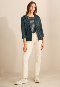 Veste zippée bleu sarcelle à manches 3/4, associée à un haut rayé bleu marine et blanc et un pantalon crème clair, le tout complété par des baskets grises.