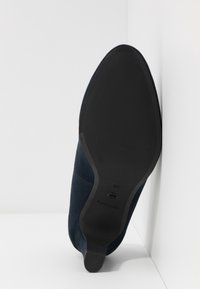 Semelle noire d'une chaussure à talon bleu marine à bout pointu, inclinée verticalement contre un mur d'angle blanc.