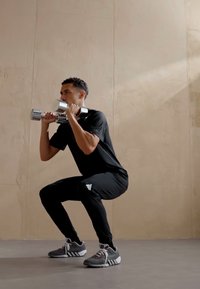 Cilvēks veic pietupienu, turot hromētus hanteles. Viņš valkā melnu krekls un bikses, kā arī pelēkus sporta apavus ar baltām akcentiem.