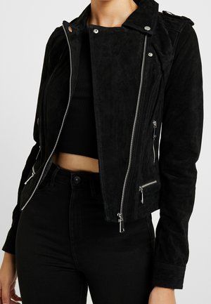 Kobieta w czarnej zamszowej kurtce motocyklowej z srebrnymi zamkami, nałożonej na czarny krótki top i czarne spodnie z wysokim stanem.
