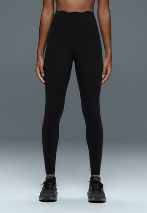Personne portant des leggings noirs taille haute et des baskets noires, debout, bras détendus le long du corps devant un fond gris uni.