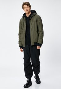 Olivenfarvet, vandafvisende jakke med hætte og forlommer. Sammen med en sort hoodie og løstsiddende sorte bukser. Sorte ankelstøvler.