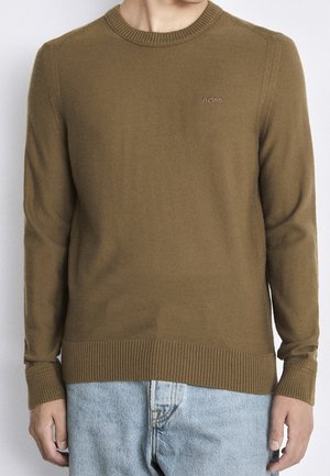 Homme portant un pull ras du cou marron avec le logo "BOSS" sur la poitrine et un jean bleu clair, debout devant un fond uni.