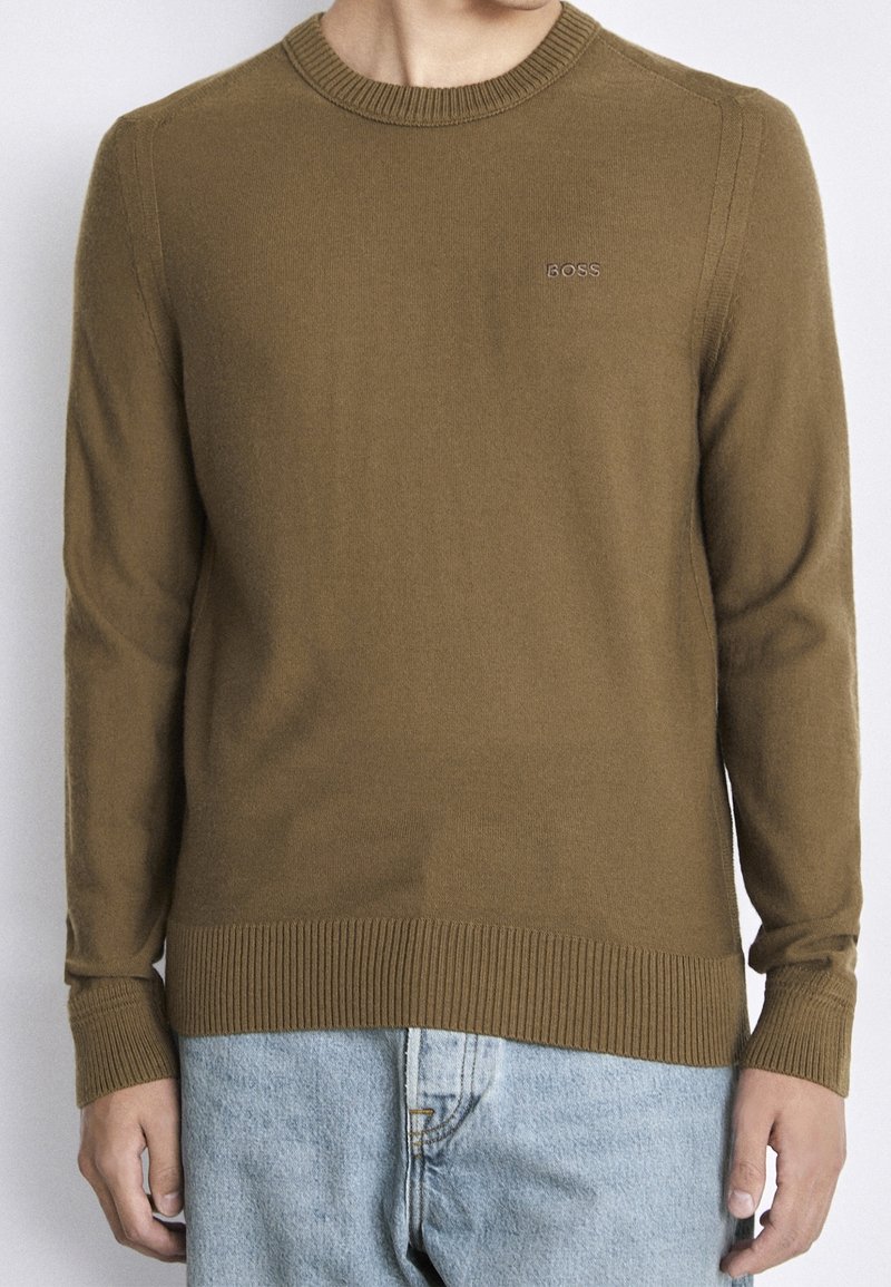 Homme portant un pull ras du cou marron avec le logo "BOSS" sur la poitrine et un jean bleu clair, debout devant un fond uni.