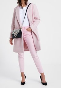 Abrigo largo de color rosa claro y textura, sobre una camisa blanca; pantalones ajustados rosa y tacones altos negros; combinado con un bolso negro con estampado de serpiente.