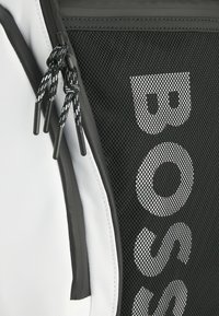Svart nät- och vit tygryggsäck med dragkedjor, med djärv text och strukturella detaljer. Accentuerad med svarta snören och klipp.