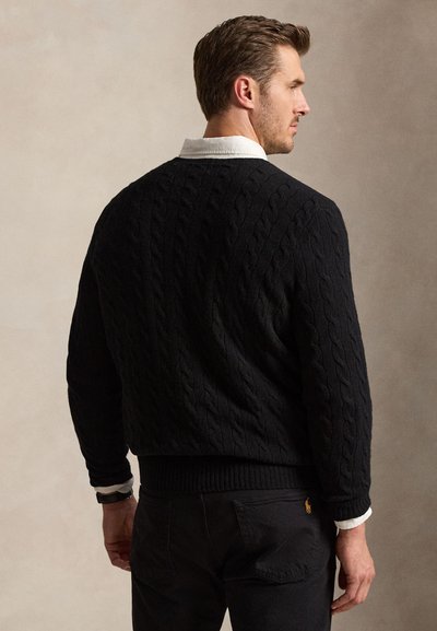 Uomo che indossa un maglione nero a trecce sopra una camicia bianca con collo, mostrato di spalle e che guarda di lato su uno sfondo semplice.