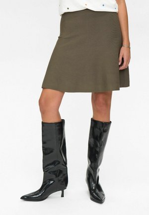 Femme portant une jupe olive de longueur genou accompagnée de bottes noires brillantes mi-mollet à talons bas sur un fond blanc.