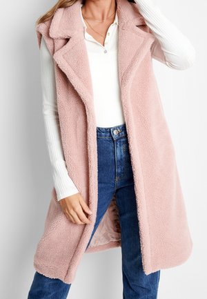 Frau trägt ärmellose pinke flauschige Weste über weißem gerippten Langarmshirt und blauer Jeans, zeigt Vorder- und Seitenansicht der Weste.