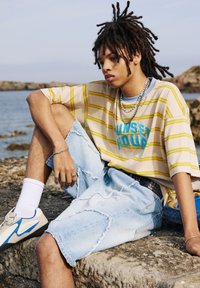 Light beige and yellow striped oversized t-shirt with "SUNSET HOUR" graphic, paired with distressed denim shorts and blue-accented sneakers.