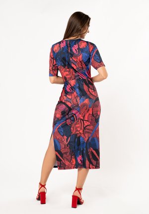 Multikleurige jurk met een bloemenpatroon in rood, blauw en zwart; korte mouwen, zijsplitsen en een getailleerde silhoët. Gecombineerd met rode blokhak sandalen.