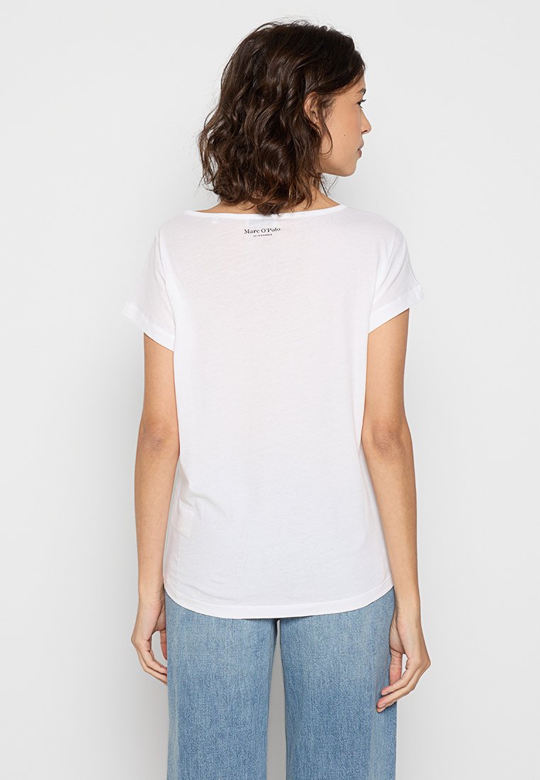 Kobieta z krótkimi, kręconymi włosami, ubrana w białą koszulkę z krótkim rękawem i niebieskie dżinsy, odwrócona plecami w stronę jednolitego białego tła.