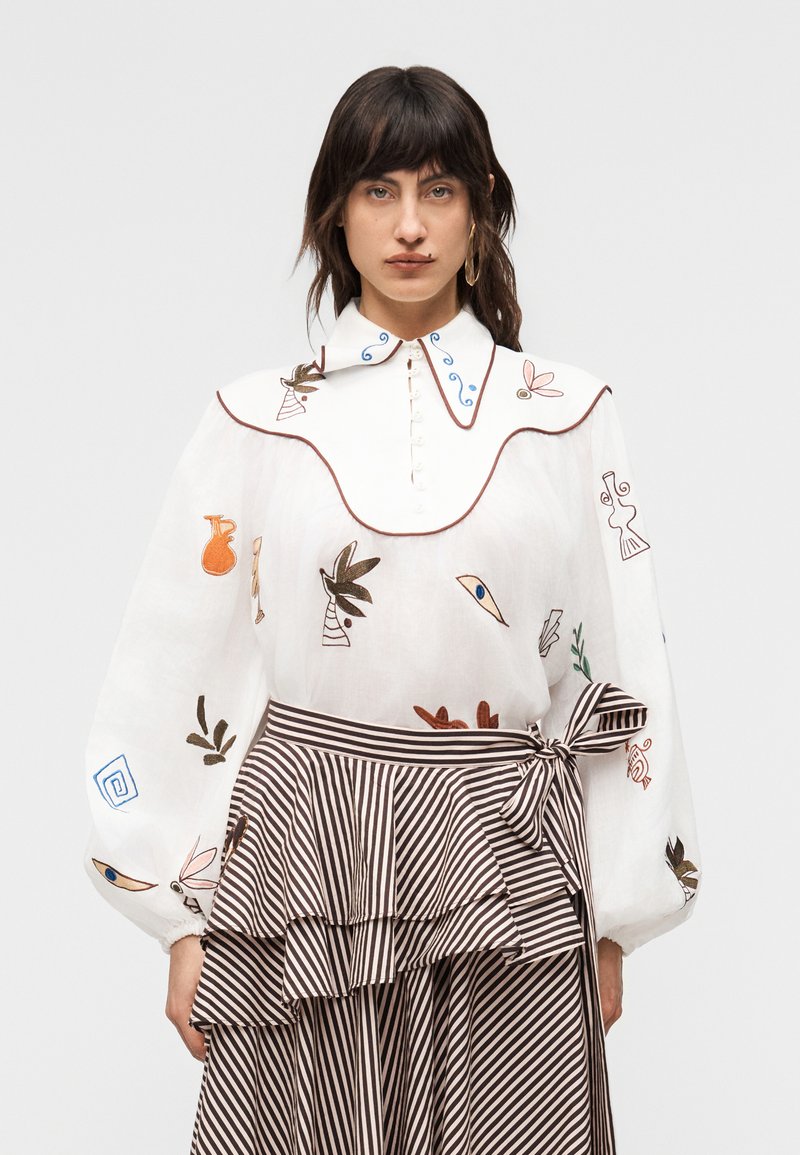 Γυναίκα που φοράει λευκό πουκάμισο με πολύχρωμη αφηρημένη κεντητική και φουσκωτά μανίκια, σε συνδυασμό με μαύρη και άσπρη ριγέ φούστα μέσης με φιόγκο.