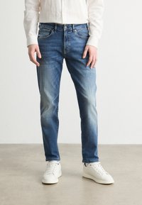 Light blue denim jeans with a slim fit, featuring light fading and whiskering details. Paired with white sneakers and a white button-up shirt.