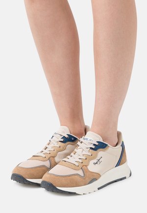 Nohy obuté v béžových, pískových a modrých volnočasových teniskách s bílými tkaničkami a silnými bílými podrážkami na bílém pozadí.