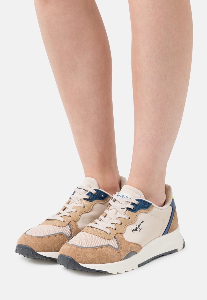 Nogi w beżowych, brązowych i niebieskich sportowych butach z białymi sznurówkami i grubymi białymi podeszwami na białym tle.