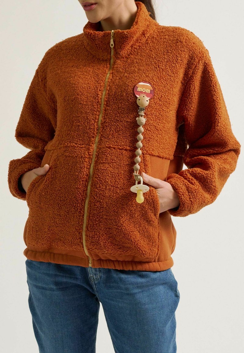 Oranger Fleece-Reißverschlussjacke mit hohem Kragen, zwei Taschen und strukturiertem Stoff; ausgestattet mit einem Clip für einen Schnuller und einem runden "Mama"-Abzeichen.