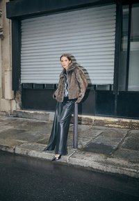 Barna műszőrme kabát magas gallérral, fekete, bőszárú bőr nadrággal és hegyes orrú fekete magassarkú cipővel, egy városi utcán.