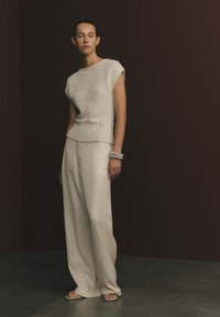 Woman wearing sleeveless textured beige top, wide-leg cream trousers, silver bracelet, and open-toe sandals standing against dark background.