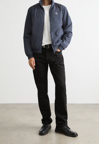 Granatowa kurtka typu bomber z wysokim kołnierzem, logo na piersi, zestawiona z białym t-shirtem, czarnymi spodniami i czarnymi butami. Gładka tekstura.