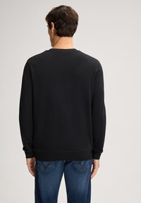 Felpa nera con collo a giro, maniche lunghe e polsini a coste. Texture liscia, design semplice, vista da dietro. Abbinata a jeans blu.