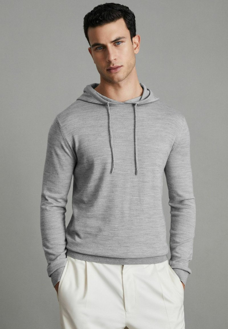 Grau gepulllter Hoodie aus weichem Stoff, mit einer Kordel, langen Ärmeln und gerippten Bündchen. Kombiniert mit hellen Hosen.