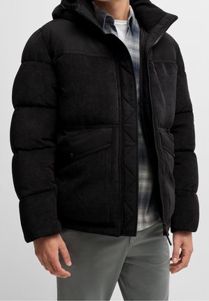 Muž v černé prošívané manšestrové bundě přes šedou a bílou kostkovanou košili a šedé kalhoty, stojící před jednobarevným pozadím.