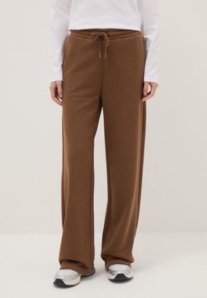 Bruine wijde sweatpants met trekkoord in de taille, gedragen met een witte top met lange mouwen en zilver-witte sneakers, op een neutrale achtergrond.