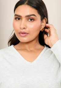 Ljust grå, långärmad topp med v-ringning. Personen bär en texturerad guldfärgad örhänge med en vågig design, hand som rör vid håret. Neutral bakgrund.