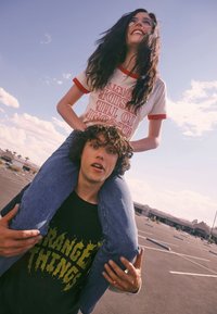 Deux jeunes adultes dans un parking extérieur. L'un porte un t-shirt blanc avec des bordures rouges et un texte, l'autre un t-shirt noir avec des illustrations jaunes.