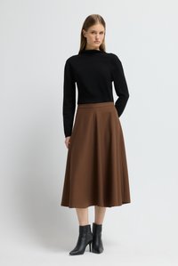 Frau steht in einem schwarzen Langarmoberteil, einem braunen Midikleid und schwarzen, hochhackigen Stiefeletten vor einem schlichten Hintergrund.