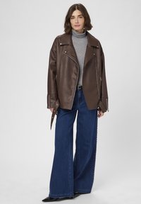 Donna che indossa una giacca di pelle marrone, dolcevita grigia, jeans blu a gamba larga con borchie laterali e scarpe nere con punta, su uno sfondo neutro.