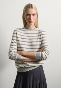 Jeune femme avec des cheveux blonds raides, portant un pull rayé crème et gris et une jupe plissée noire, se tenant les bras croisés contre un mur blanc.
