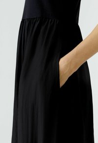 Schwarzes Kleid aus leichtem Stoff mit tailliertem Oberteil und fließendem, gerafftem Rock. Verfügt über seitliche Taschen und eine glatte Textur.