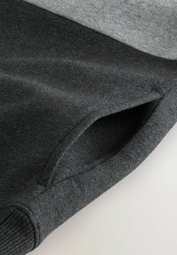 Nahaufnahme eines dunkelgrauen Stoffärmels mit Seitentasche und Rippbündchen, das Textur- und Nahtdetails auf hellem Hintergrund zeigt.