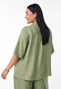 Blusa verde oliva realizzata in tessuto testa di moro, con una vestibilità morbida, maniche corte e un dettaglio nella parte posteriore.