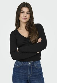 Top nero a maniche lunghe con scollatura a V, caratterizzato da un design arricciato sul davanti. Abbinato a jeans in denim blu a vita alta.