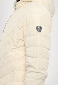 Lys beige polstret jakke med horisontalt quiltet mønster, med hette og en liten svart logo-patch på venstre skulder.