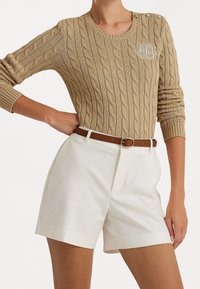 Beige kabelstickad tröja med rund halsringning och dekorativa knappar, kombinerad med vita skräddarsydda shorts och ett brunt läderbälte.