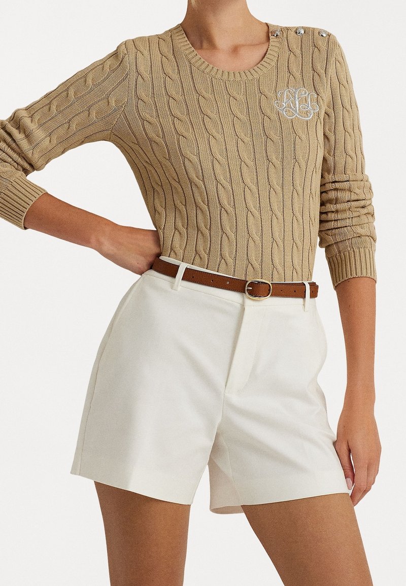 Beige kabelstickad tröja med rund halsringning och dekorativa knappar, kombinerad med vita skräddarsydda shorts och ett brunt läderbälte.