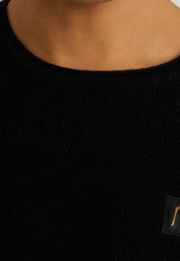 Svart stickad tröja med en strukturerad yta och rund halsringning, med en liten, gul varumärkesetikett på nedersta högra sidan.