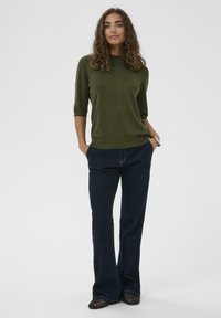 Maglione corto a maniche corte verde oliva abbinato a jeans a zampa d'elefante blu scuro. Il maglione ha una finitura testurizzata; i jeans presentano cuciture a vista.