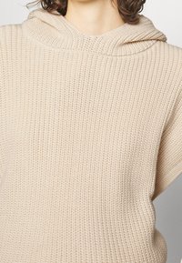 Maglione beige in maglia con un ampio cappuccio morbido e texture a costine. Presenta maniche corte e una vestibilità rilassata, adatto per un look casual.
