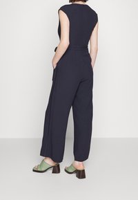 Žena v černém overal s bezrukávem a páskem, stojící s rukama v kapsách, v zelených sandálech na podpatku na bílém pozadí.