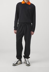 Maglietta polo nera a maniche lunghe con colletto arancione e logo ricamato, abbinata a pantaloni sportivi neri con cordino e sneakers bianco-neri.