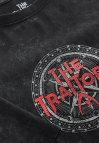 Zwarte katoenen T-shirt met een cirkelvormig grafisch ontwerp en de tekst "DE VERADER" in vette rode letters met gedetailleerde accenten.