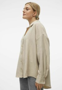 Camicia beige con bottoni realizzata in un tessuto texturizzato, vestibilità relaxed e spalle scese. Presenta un colletto e maniche lunghe. Abbinata a pantaloni grigi.