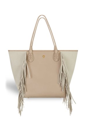 Tan leather tote bag with beige canvas sides, featuring long, neutral fringes, braided handles, and a round gold accent on the front.